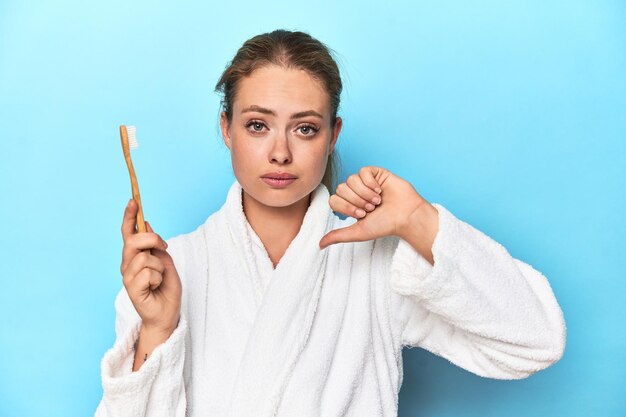 Photo blonde in bathrobe with a toothbrush showing a dislike gesture thumbs down disagreement concept