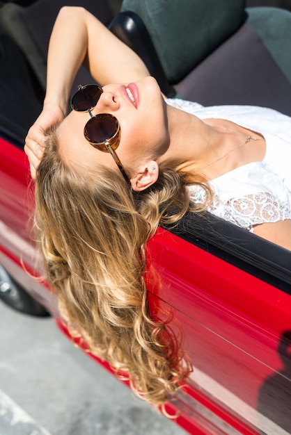 海で赤い車に座っているサングラスのブロンドの美しい女性。海の眺め。休暇の概念。幸せ。自由。美しい晴れた夏の日のロードトリップ