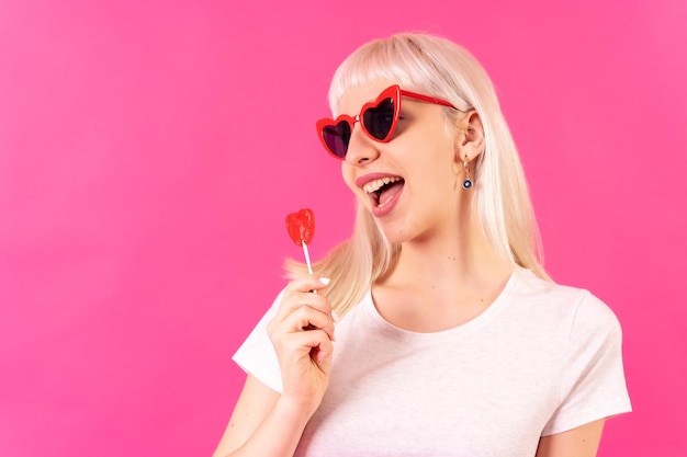 Blonde blanke meisje in studio op roze achtergrond glimlachend in glazen harten met een lolly