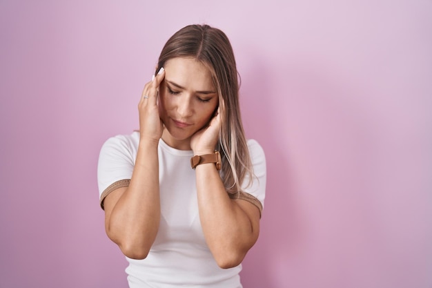 Blonde blanke vrouw die over roze achtergrond staat met hand op hoofdhoofdpijn omdat stress lijdt aan migraine