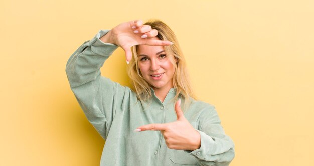 Blonde blanke vrouw voelt zich gelukkig, vriendelijk en positief glimlachend en maakt een portret of fotolijst met handen
