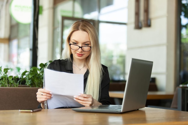 Blonde blanke zakenvrouw onderzoekt vellen documenten en zit aan een tafel met een laptop in een openluchtcafé Moderne zakenvrouw Lifestyle