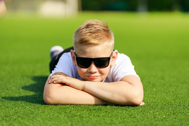 A blonde boy in dark glasses lies on a football field. High quality photo