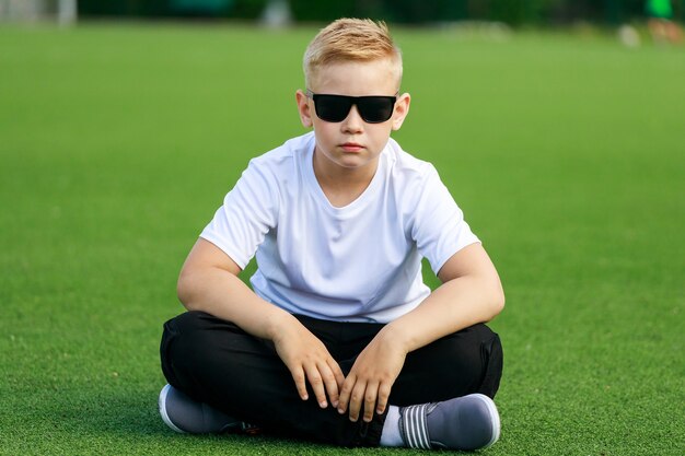 A blonde boy in dark glasses sitting on a football field in the summer. High quality photo