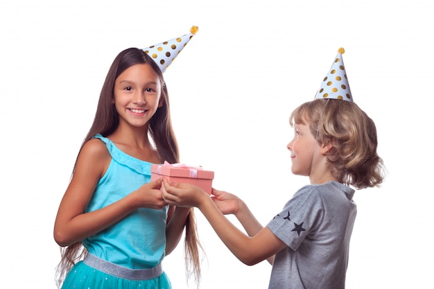 Blonde boy in festive paper hat gives present in gift box to happy girl on her birthday party