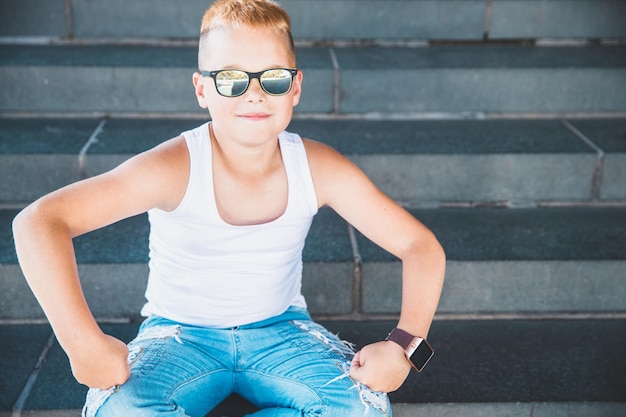 Photo blonde boy in jeans and white t-shirt sits on the steps