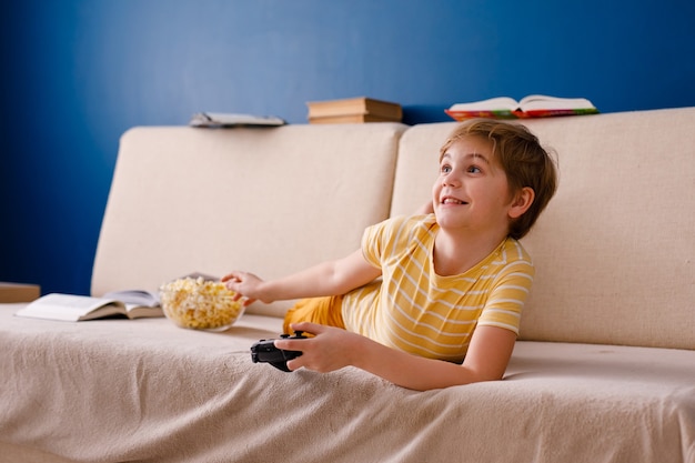 Blonde boy plays with gamepad instead of lessons and eats popcorn.