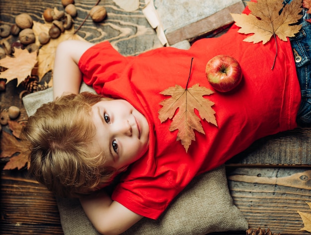 Il ragazzo biondo che riposa con una mela sullo stomaco si trova sul pavimento di legno in foglie di autunno. i più grandi sconti per tutti i vestiti autunnali per bambini. bambino che gioca in autunno.