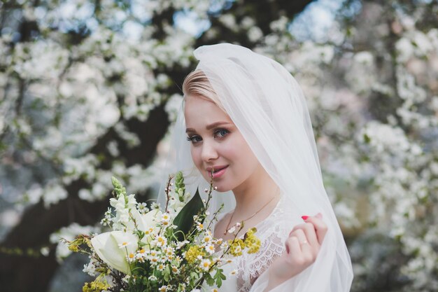 La sposa bionda tiene un bouquet da sposa nel parco di primavera, abito da sposa