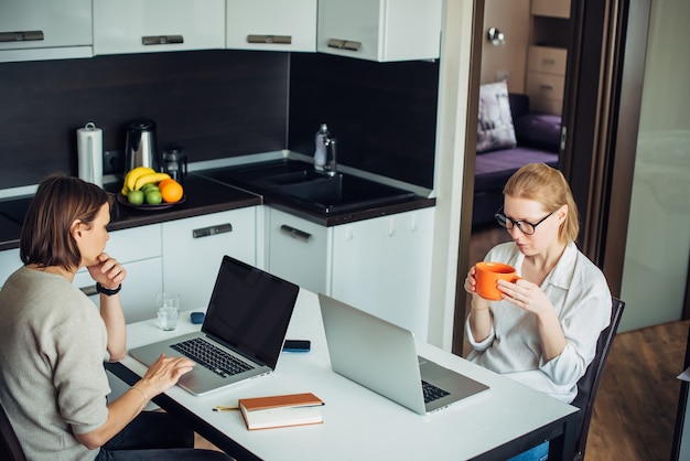 Blonde and brunette work on laptops, sitting at a table in the kitchen facing each other. Coworking in cozy home interior.