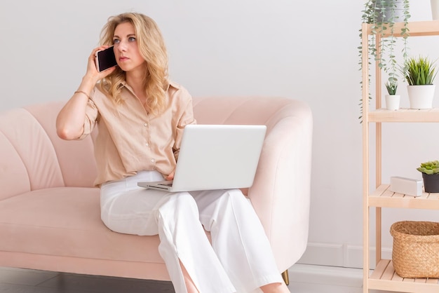 A blonde business woman with a gray open laptop on her knees calls on a pink smartphone