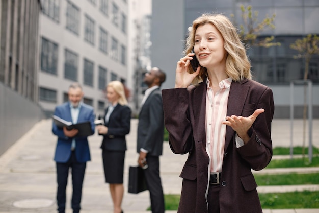 Блондинка-бизнесвумен стоит и разговаривает по телефону перед современным офисом