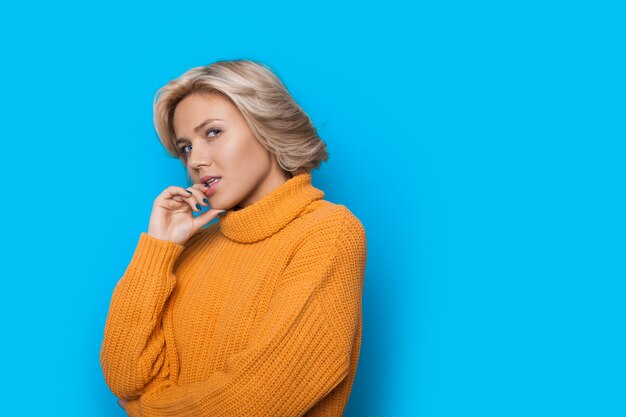 Blonde caucasian charming woman in a yellow sweater touching lips and posing on a blue wall with free space