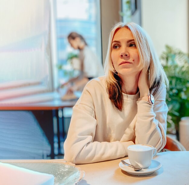 A blonde caucasian joyful girl in a light sweater sits in a restaurant near the window at a table