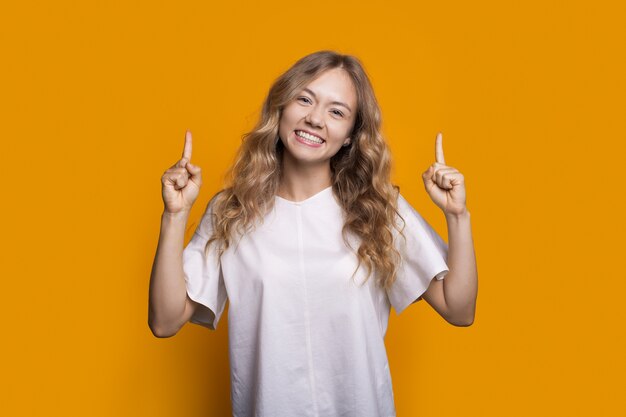 Blonde caucasian lady in a white t-shirt is smiling toothy and pointing up on a yellow studio wall advertising something