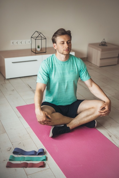Blonde caucasian man practice yoga at home on a pink carpet on the floor with some elastic bands near