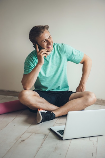 Uomo caucasico biondo che parla sul telefono prima di fare esercizi di stretching sul pavimento con un computer portatile