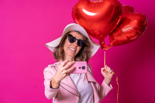 A blonde caucasian woman having fun with a white hat and sunglasses in a nightclub with some heart balloons taking a selfie with her mobile phone pink background