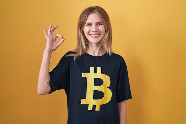 Blonde caucasian woman wearing bitcoin t shirt smiling positive doing ok sign with hand and fingers. successful expression.