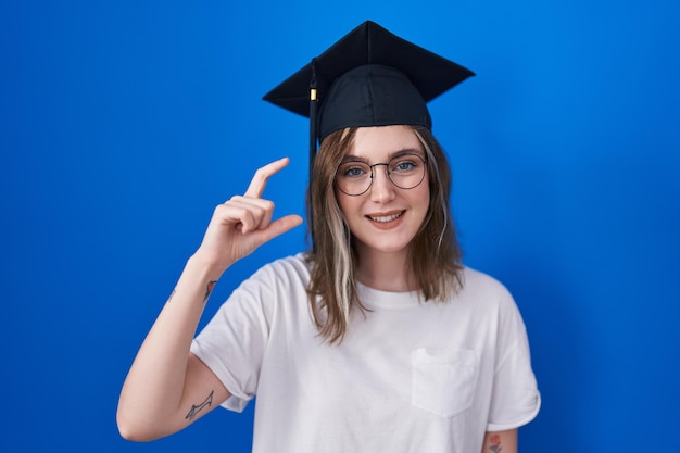 Blonde caucasian woman wearing graduation cap smiling and confident gesturing with hand doing small size sign with fingers looking and the camera measure concept