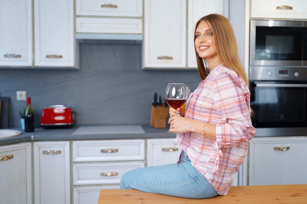 Donna caucasica bionda con un bicchiere di vino rosso in piedi nella sua cucina