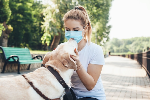 Donna caucasica bionda con mascherina medica sul viso che cammina nel parco con un labrador baciandolo e abbracciandolo