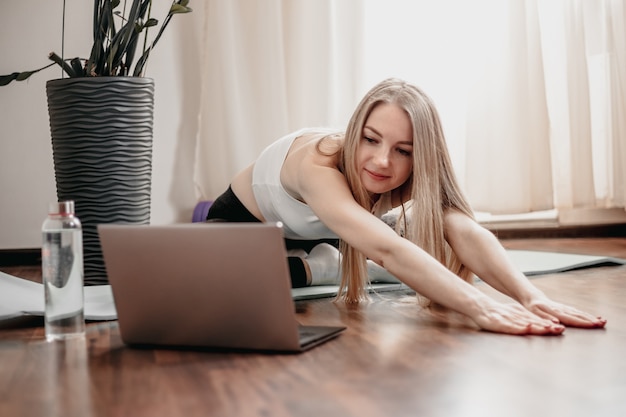 Blonde caucasian young woman doing pilates at home online and looking at the laptop screen and smile