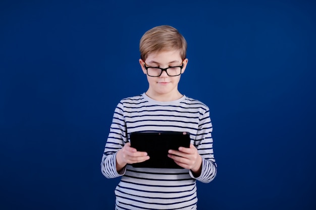 Blonde child boy with big glasses playing with tablet  on blue wall. Education concept.