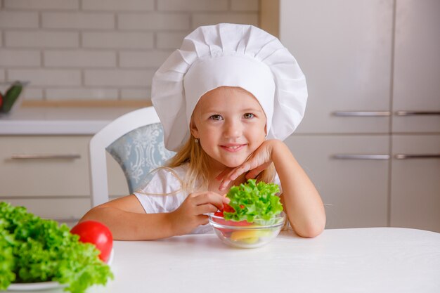 белокурый ребенок в шляпе шеф-повара на кухне ест овощи