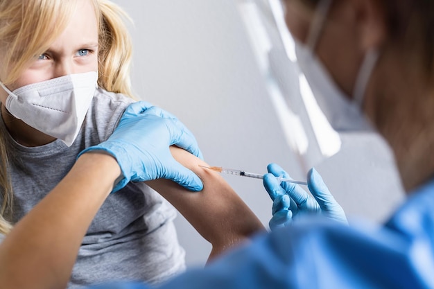 Blonde child girl looking at the doctor with syringe in hand and refuses syringe with vaccine while being vaccinated or receiving a shot during covid19 pandemic