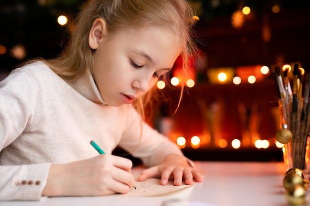 Lettera di scrittura della ragazza del bambino biondo a santa claus o disegnare qualcosa sul con bokeh giallo caldo