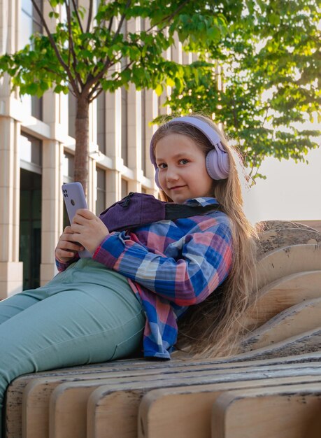 Blonde child in headphones listening to music podcast smartphone outside Young girl learn online lesson enjoy audio book