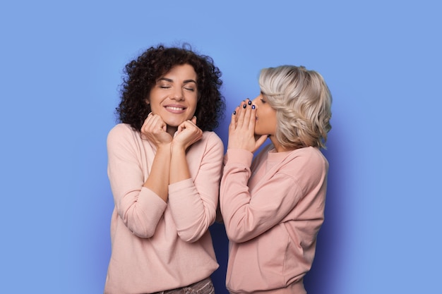 Blonde curly woman is whispering something to her brunette sister smiling on a blue wall