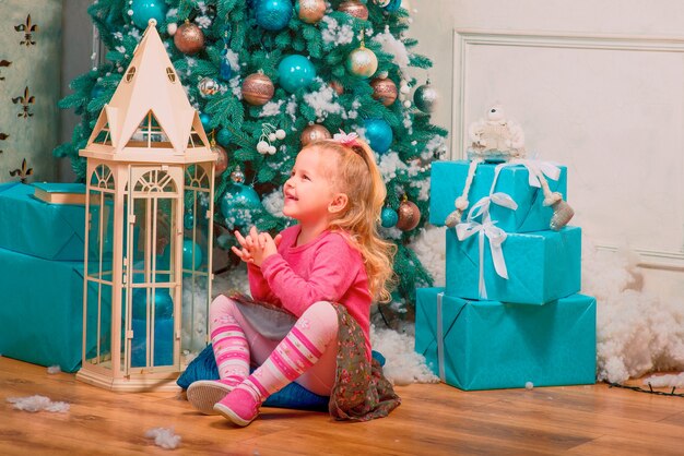 Blonde cute little girl sitting and smiling nearly decorated christmas tree and new year presents