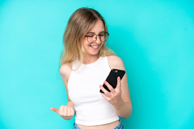 Blonde english young girl isolated on blue background using\
mobile phone and doing victory gesture