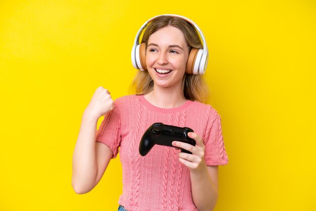 Blonde english young girl playing with a video game controller isolated on yellow background celebrating a victory