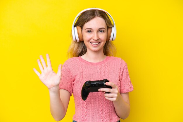 Blonde english young girl playing with a video game controller isolated on yellow background counting five with fingers