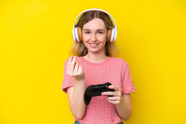 Blonde english young girl playing with a video game controller isolated on yellow background making money gesture