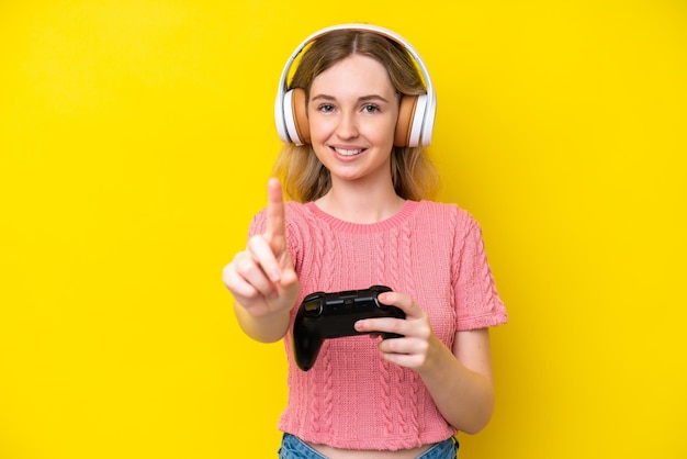 Blonde English young girl playing with a video game controller isolated on yellow background showing and lifting a finger