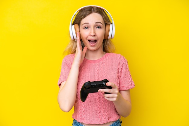 Blonde English young girl playing with a video game controller isolated on yellow background with surprise and shocked facial expression