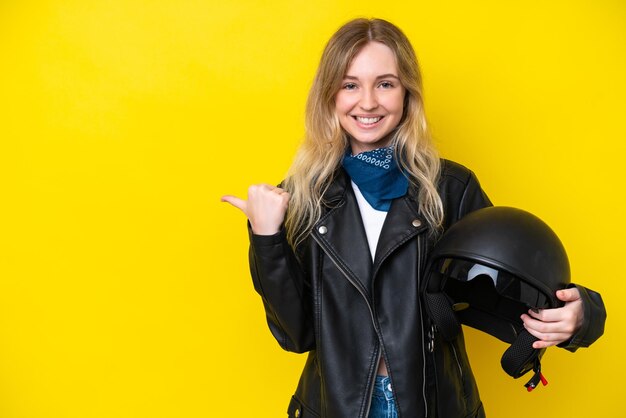 Blonde english young girl with a motorcycle helmet isolated on yellow background pointing to the side to present a product