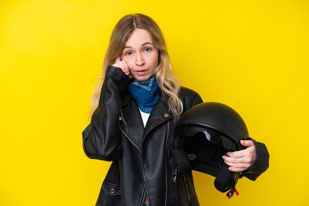 アイデアを考えて黄色の背景に分離されたオートバイのヘルメットを持つ金髪の英語の若い女の子