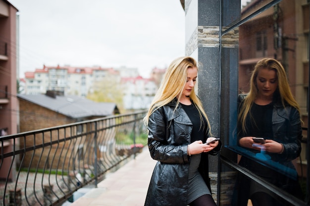 Blonde fashionable girl in long black leather coat posed against large window of building.