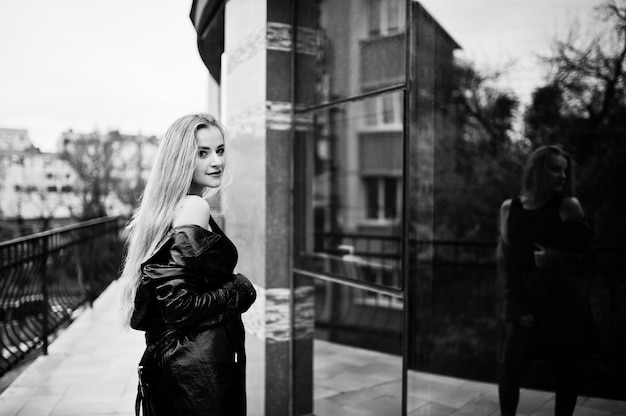 Blonde fashionable girl in long black leather coat posed against large window of building.