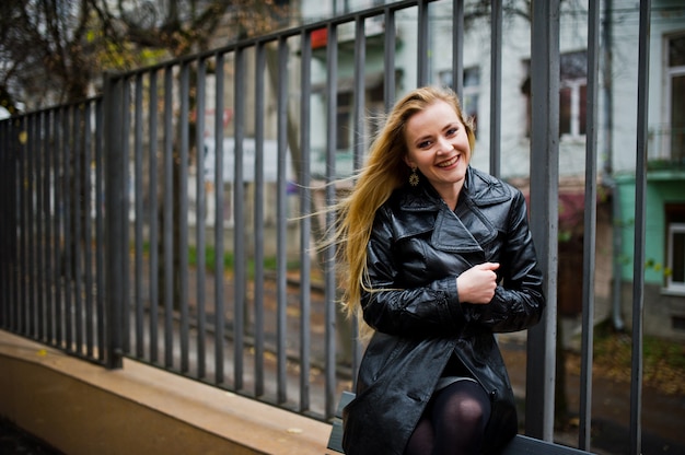 Blonde fashionable girl in long black leather coat posed on bench.