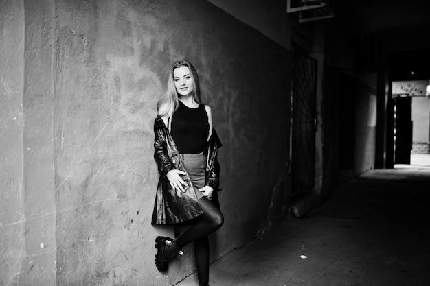 Blonde fashionable girl in long black leather coat posed near street tonel with orange wall