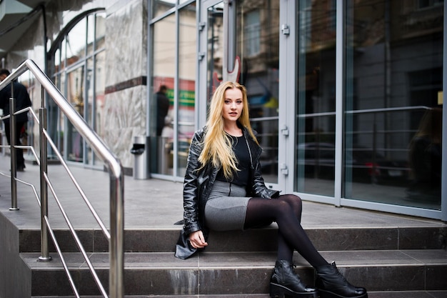 Blonde fashionable girl in long black leather coat posed at stairs