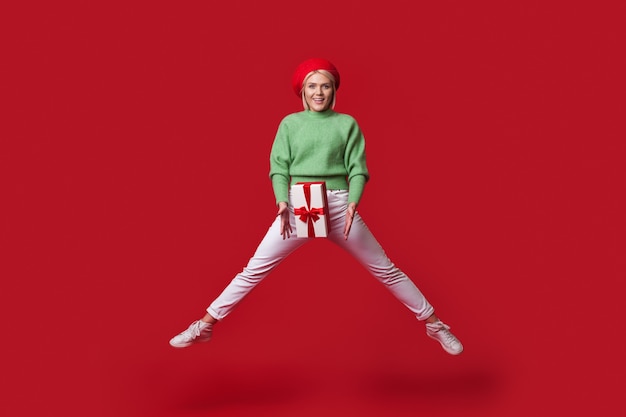 Blonde fashionable woman is jumping on a red wall holding a present box and wear a hat smiling at camera