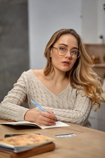 Blonde female blogger in glasses beige sweater sitting in kitchen at wooden table going to write som...