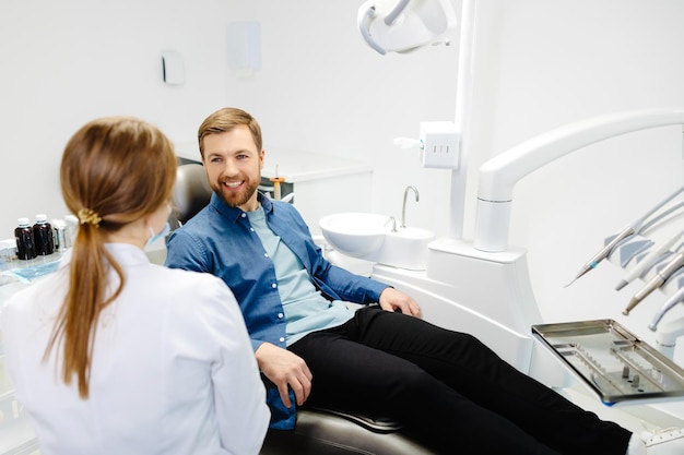 Blonde female dentist in dental office talking with male patient and preparing for treatment Handsome bearded man in dentist chair looking at his doctor with smile